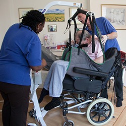 A brain injured man who has his care funded, is helped into a wheelchair by two carers