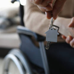 A person in a wheelchair shakes hands with someone handing over the keys to their home illustrating the home adaptations that can be made after serious injury
